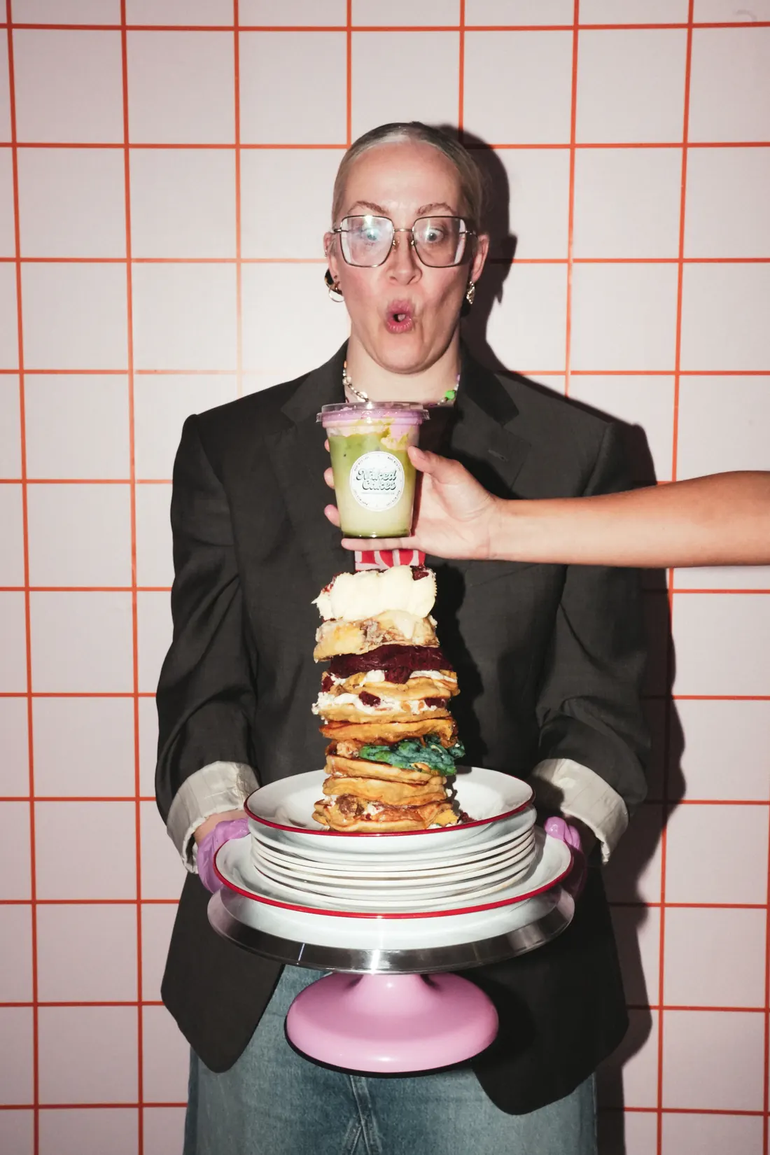 Jessie Van Osselt met een hoge toren van pancakes en cookies, voor een tegelmuur
